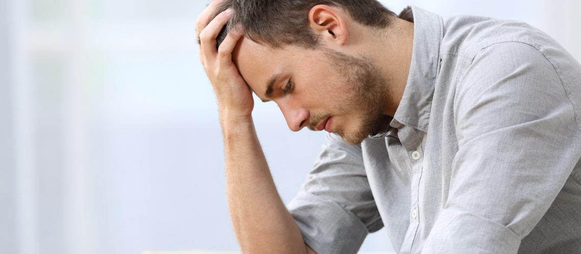 Side view of a sad man with a hand on the head sitting on a couch in the living room at home